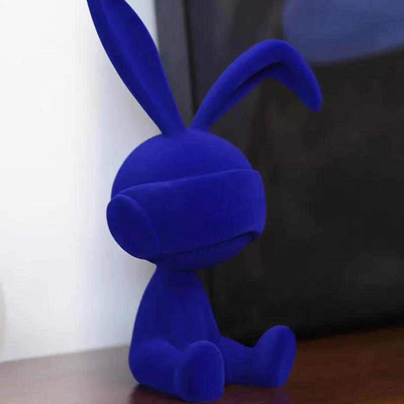 Small resin rabbit ornament wearing a suede blindfold, shown in blue, green, and red colour options.