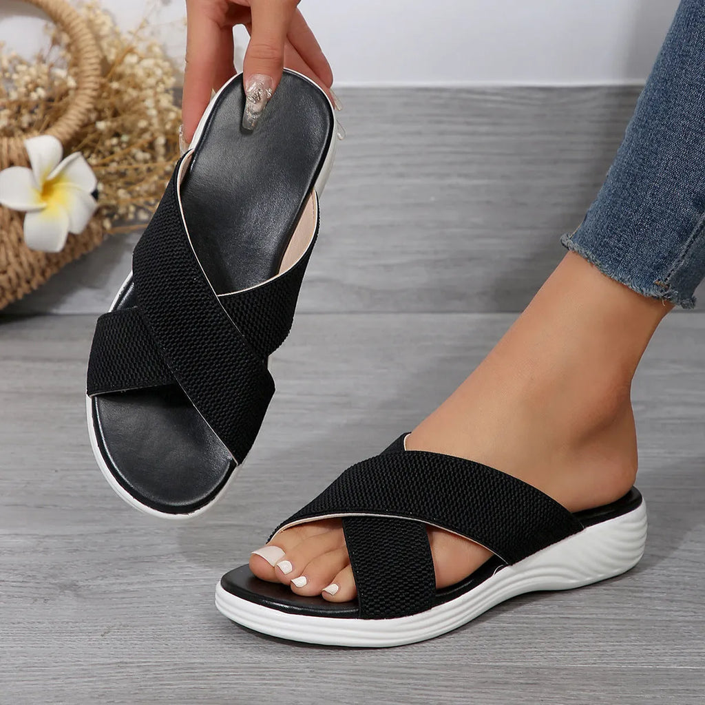 Women's black peep-toe wedge slippers with a cross strap and thick rubber sole on a grey background.