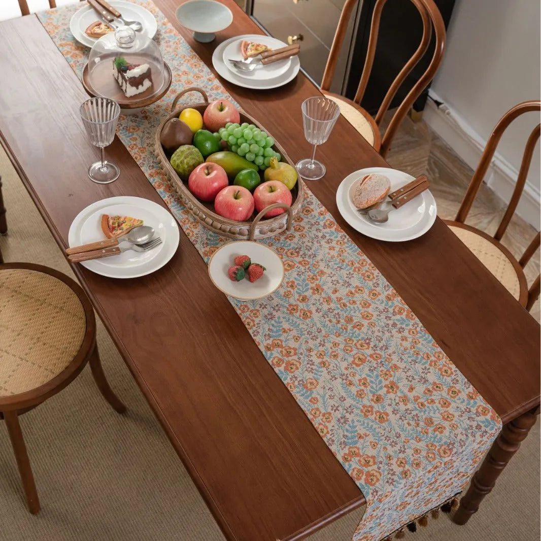 Beige Jacquard table runner with floral leaf pattern and long tassel fringe on a wooden table.