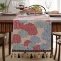 Beige Jacquard table runner with floral leaf pattern and long tassel fringe on a wooden table.