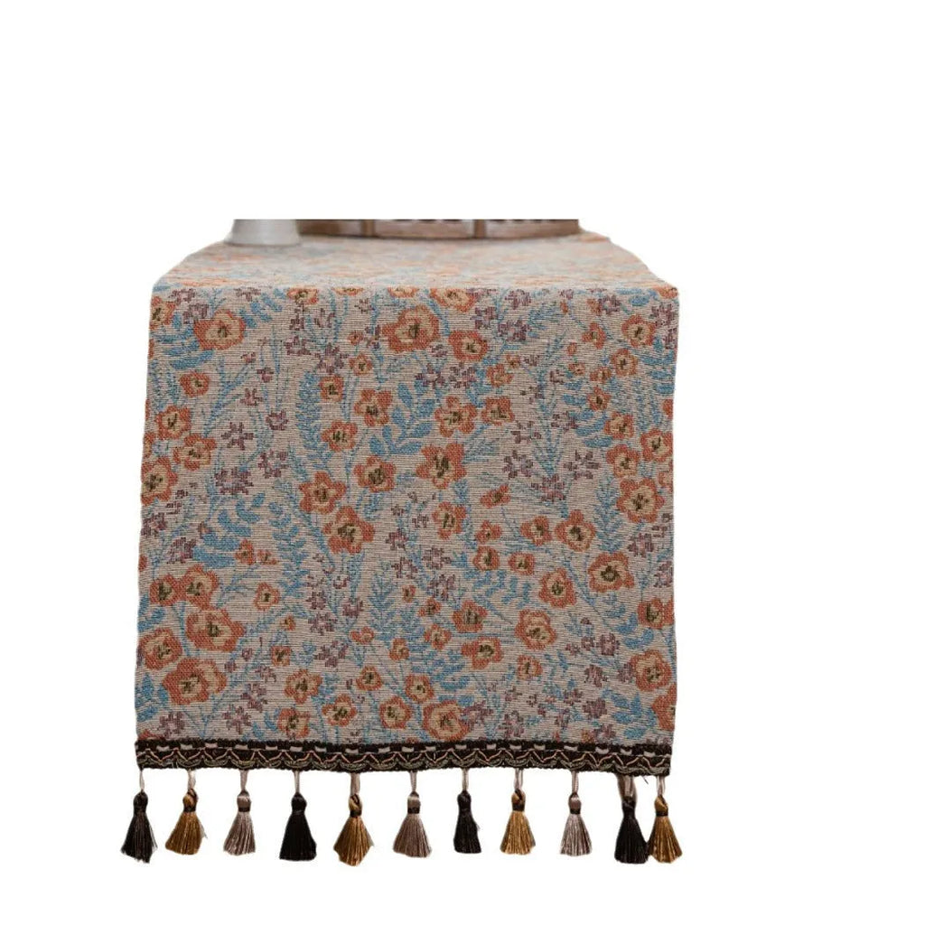 Beige Jacquard table runner with floral leaf pattern and long tassel fringe on a wooden table.