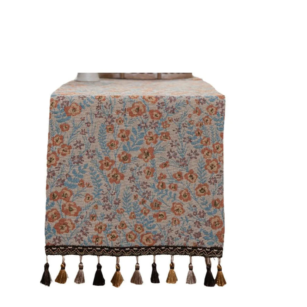 Beige Jacquard table runner with floral leaf pattern and long tassel fringe on a wooden table.