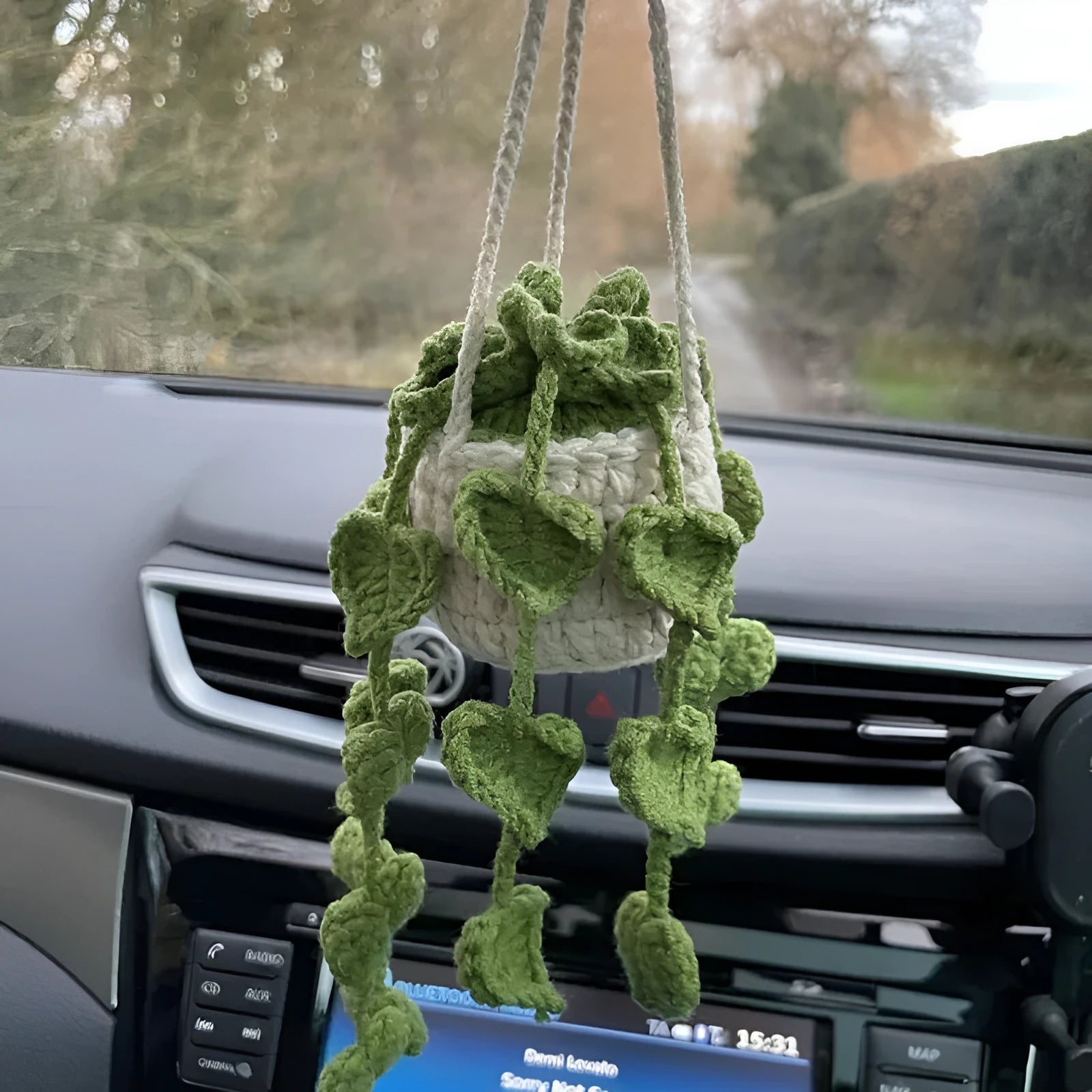 Green and white crocheted car plant decoration with hanging rope on a car dashboard.