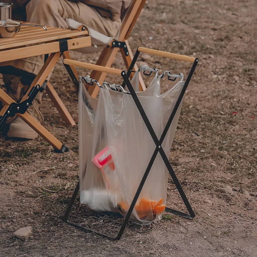 Black iron folding trash rack with wooden handle holding a bin bag outdoors.