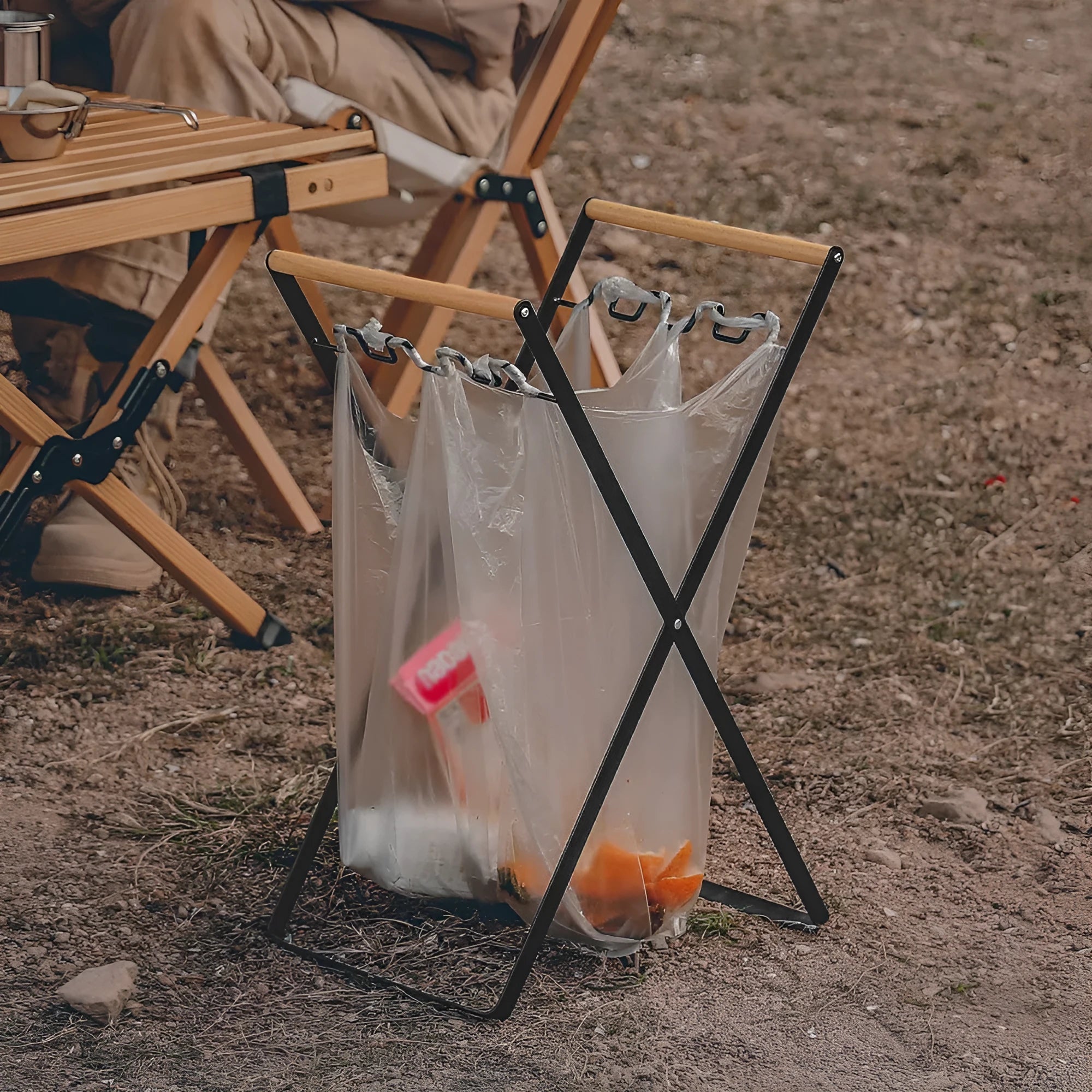 Black iron folding trash rack with wooden handle holding a bin bag outdoors.