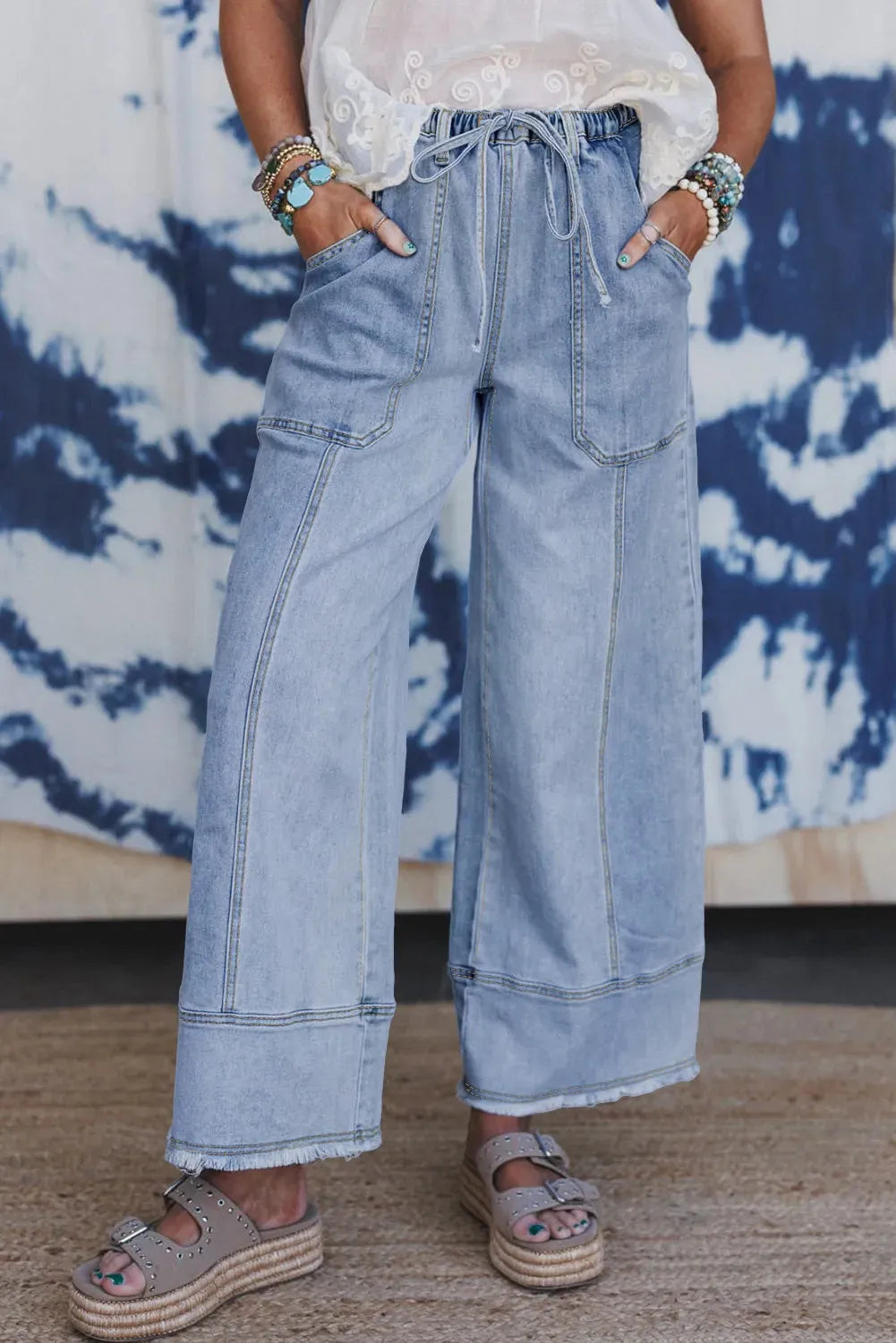 Beau blue wide leg denim trousers with a drawstring waist and raw hem detail laid flat.