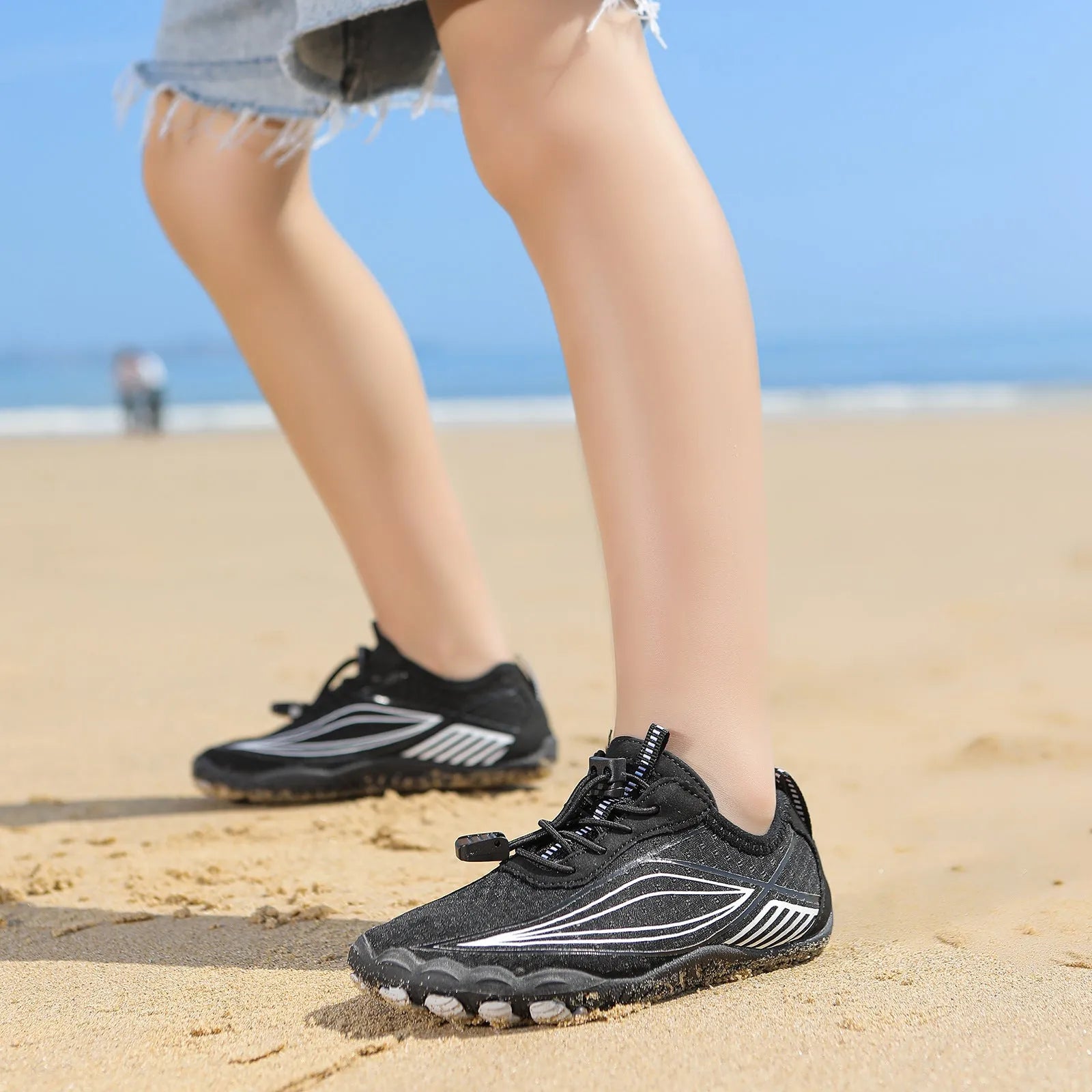 Children's elastic band water shoes in blue with a non-slip rubber sole, shown on a sandy beach.